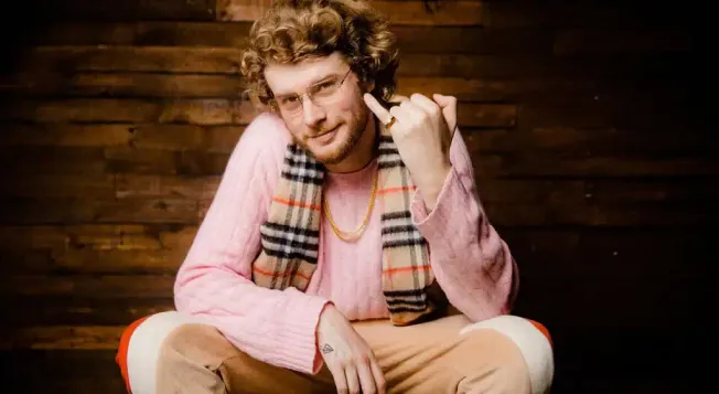 Portrait of young man with curly hair wearing pink sweater and glasses, sitting on floor in front of wooden wall, making a re