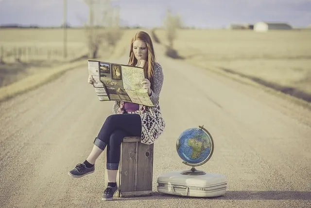 A young woman sits on a wooden crate on an empty rural road, reading a newspaper with a globe and suitcase beside her, repres