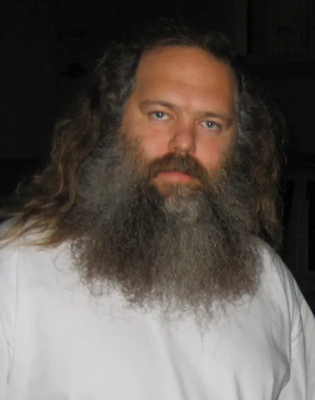 Long-haired man with a full beard and serious expression in a casual white shirt, indoor setting.