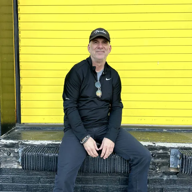 Bright yellow wall with black horizontal lines and a man in black Nike sportswear sitting on a weathered wooden bench, casual