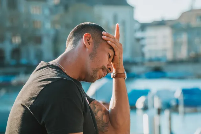A man with tattoos in distress holding his forehead near a harbor with boats, illustrating stress or worry in an urban outdoo