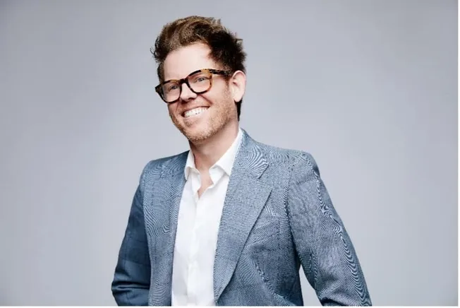 Fashionable man in gray blazer with glasses smiling in studio portrait - Hypebot.