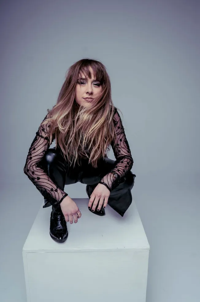 Elegant woman with long wavy hair wearing a black lace top and leather pants, seated on a white pedestal against a neutral ba