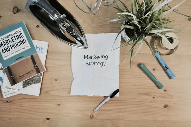 Marketing strategy planning on wooden desk with books, markers, iron, plant, and crumpled paper.