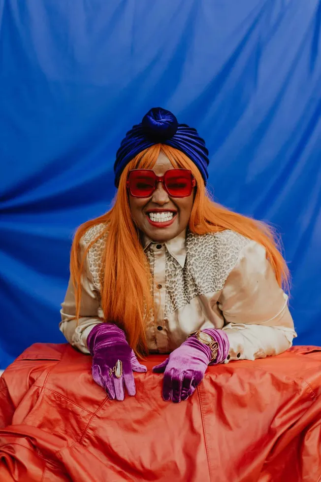 Colorful fashionable woman with bright orange hair, oversized red sunglasses, purple velvet gloves, bold accessories, stylish