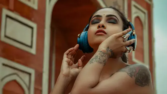 Headshot of a woman with tattoos enjoying music with blue headphones, standing outdoors against a historic brick building, ca