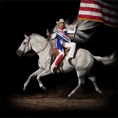 A female rodeo rider in patriotic attire on a white horse holding an American flag during a rodeo event.