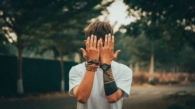 Young man covering his face with hands outdoors, showcasing lifestyle and expressive pose.