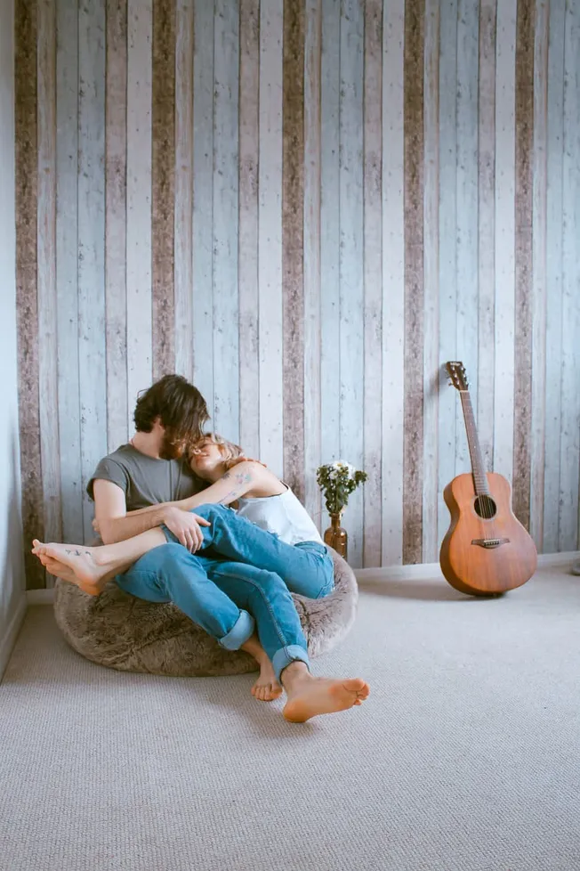 A couple cuddling on a cozy bean bag with an acoustic guitar beside them in a room with rustic wooden paneling, capturing int