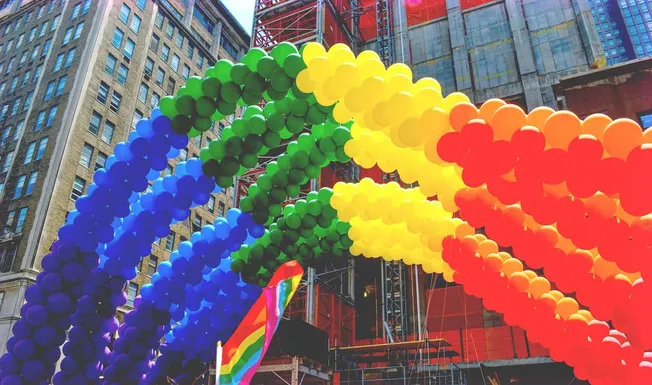 Vibrant rainbow balloons arching over a city street during a Pride parade, with colorful buildings and a rainbow flag in the