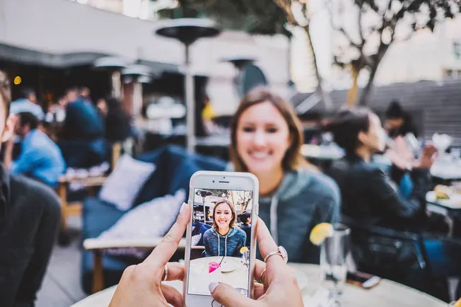 Woman taking a photo of a smiling woman at an outdoor restaurant or cafe, capturing a joyful moment with smartphone photograp