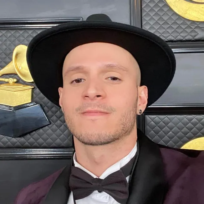 Stylish man in a black hat and tuxedo at a music award show, standing in front of a backdrop with gold gramophone awards and