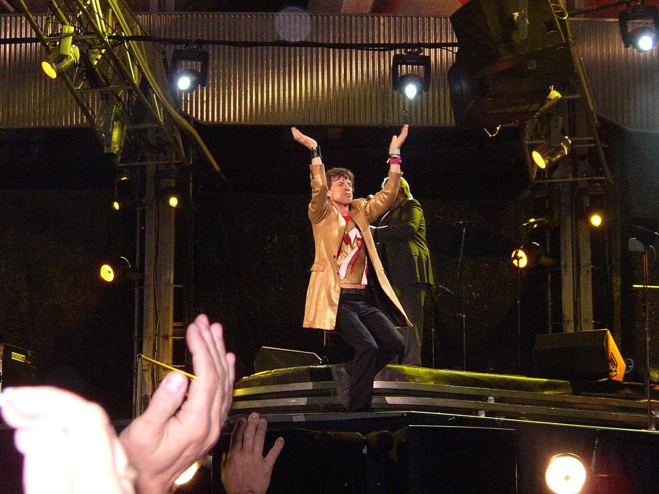 Vibrant live performance scene featuring a male singer dancing on stage under colorful lighting, with an enthusiastic audience clapping in front. The stage has professional lighting and sound setup, capturing the energetic atmosphere of a concert.