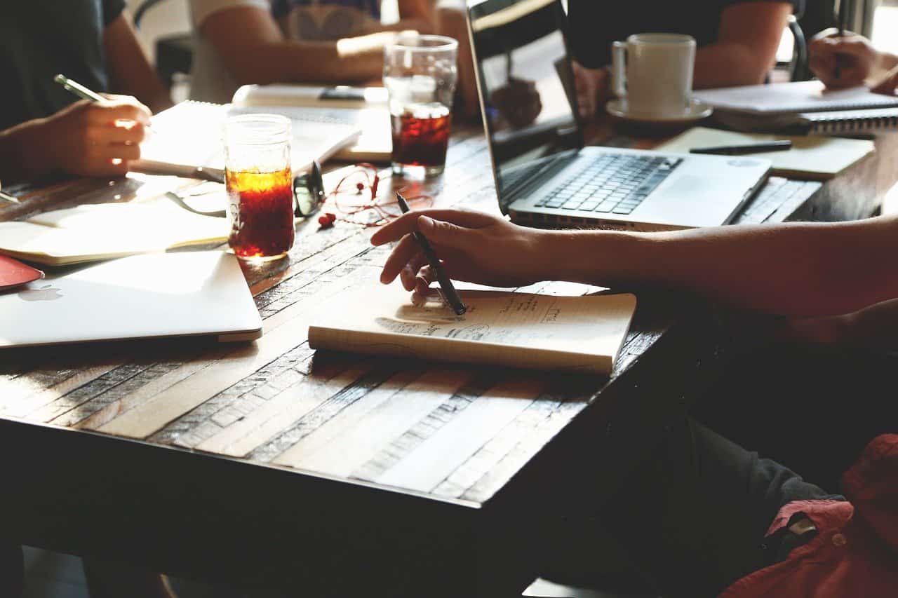Focused collaboration meeting at a wooden table with laptops, notebooks, and beverages, emphasizing teamwork, productivity, and creative brainstorming in a modern workspace.
