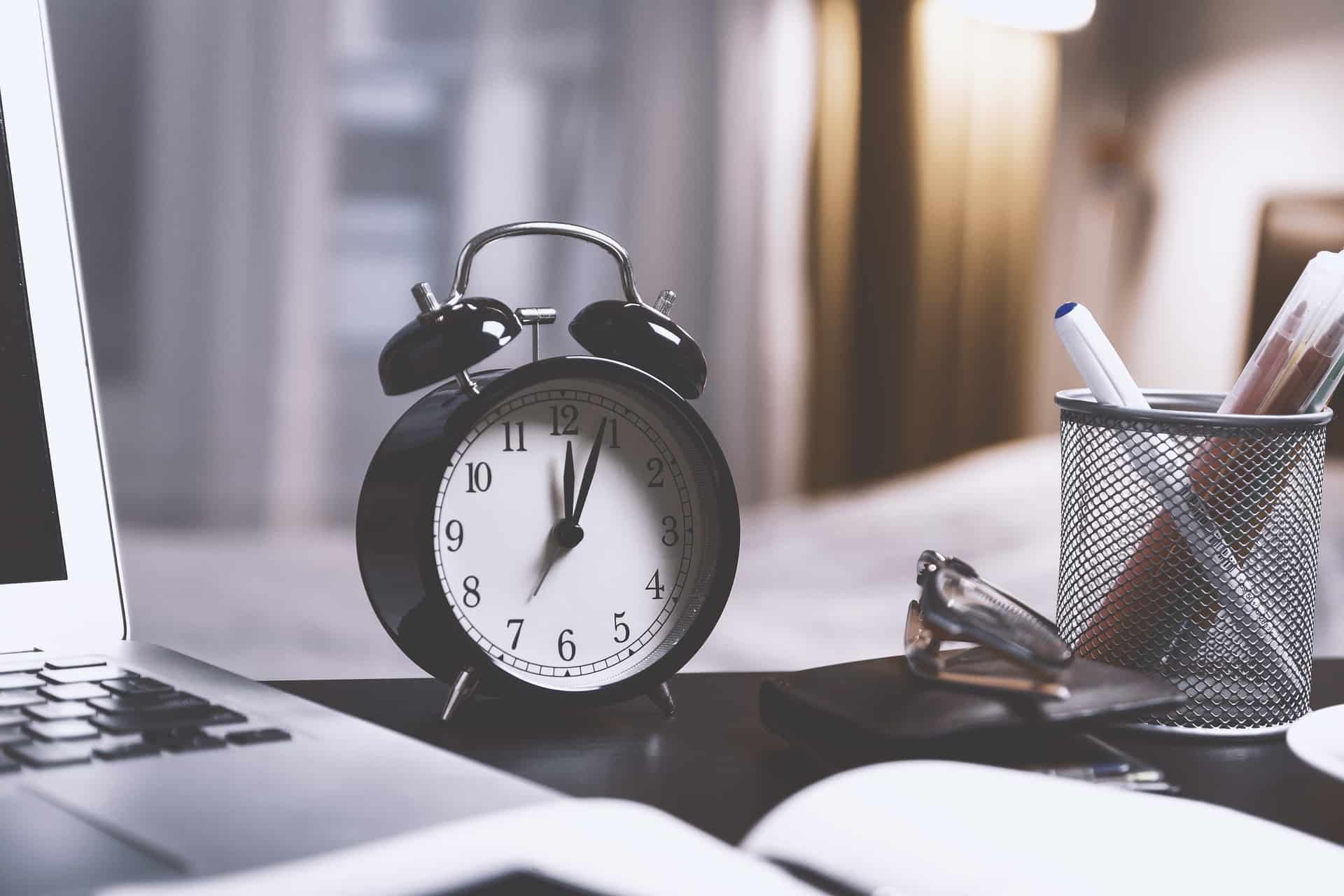 Alarm clock on a desk with a laptop, pen holder, and glasses, symbolizing productivity and time management in the workspace.