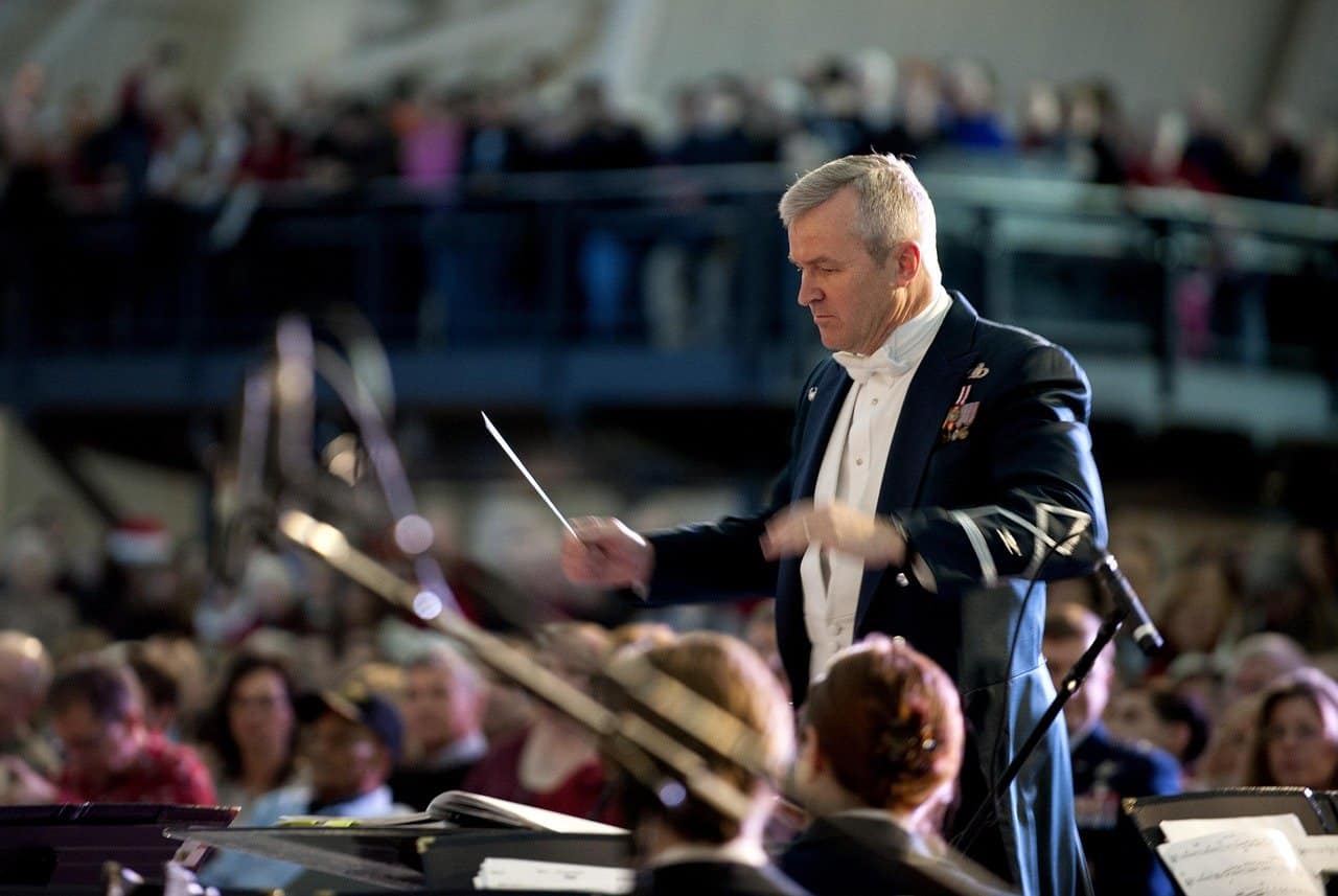 Orchestra conductor leading a symphony in a concert hall, dressed in a formal tuxedo with medals, guiding musicians during a live performance, with an engaged audience in the background.