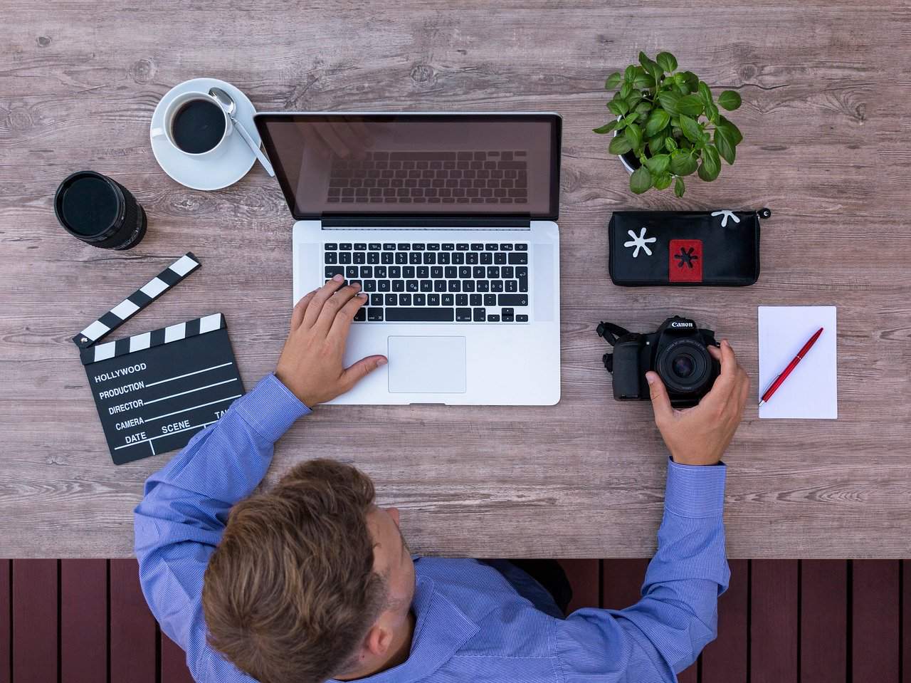 Aesthetic flat lay of a person working with a laptop, camera, clapperboard, and photography accessories on wooden desk, creative workspace for film, video production, and digital media content creation.