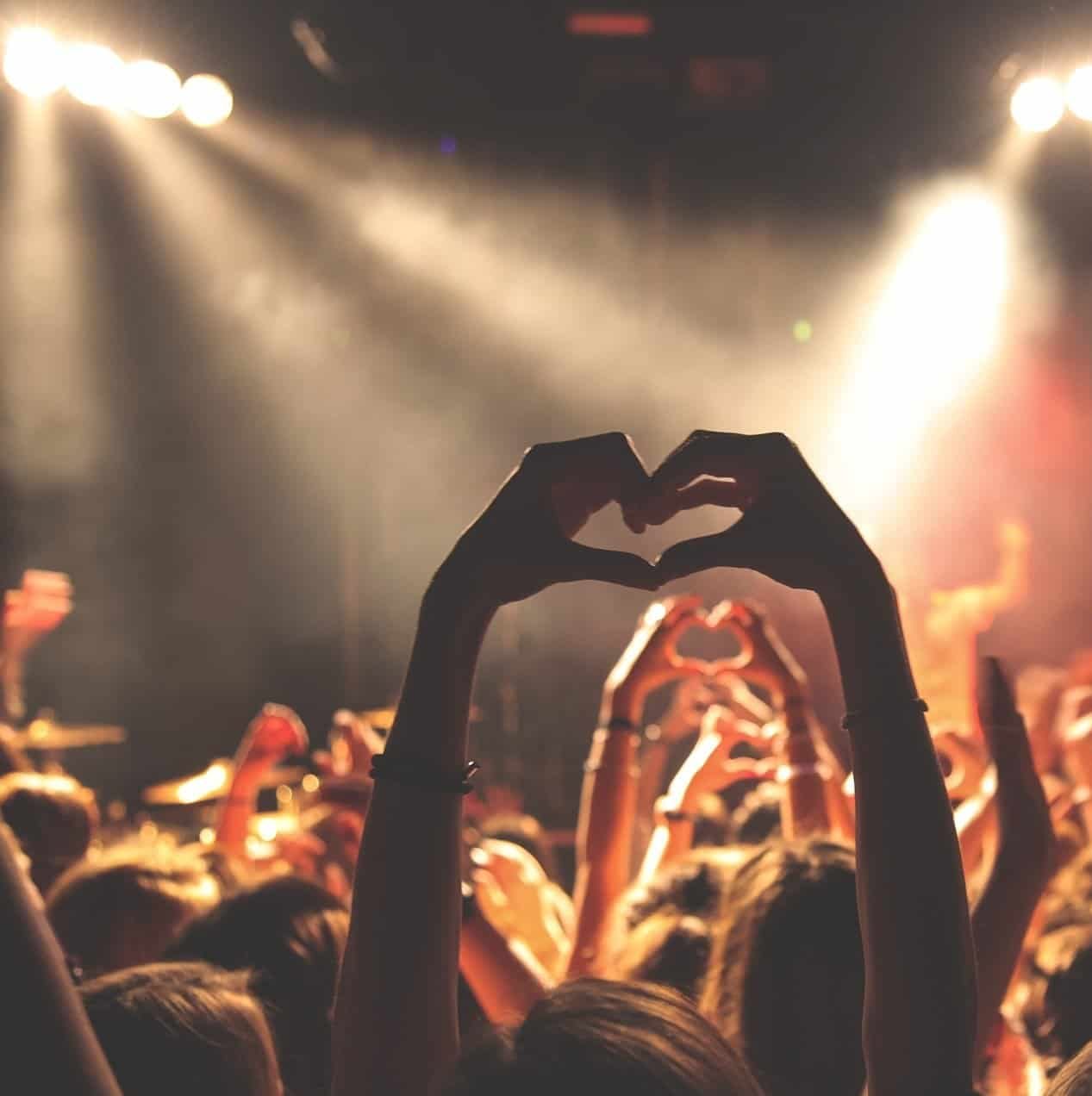 Heart shape hand gesture at concert or music festival, lively crowd with hands raised, stage lighting, music fans enjoying live performance, energetic atmosphere for entertainment and music events.