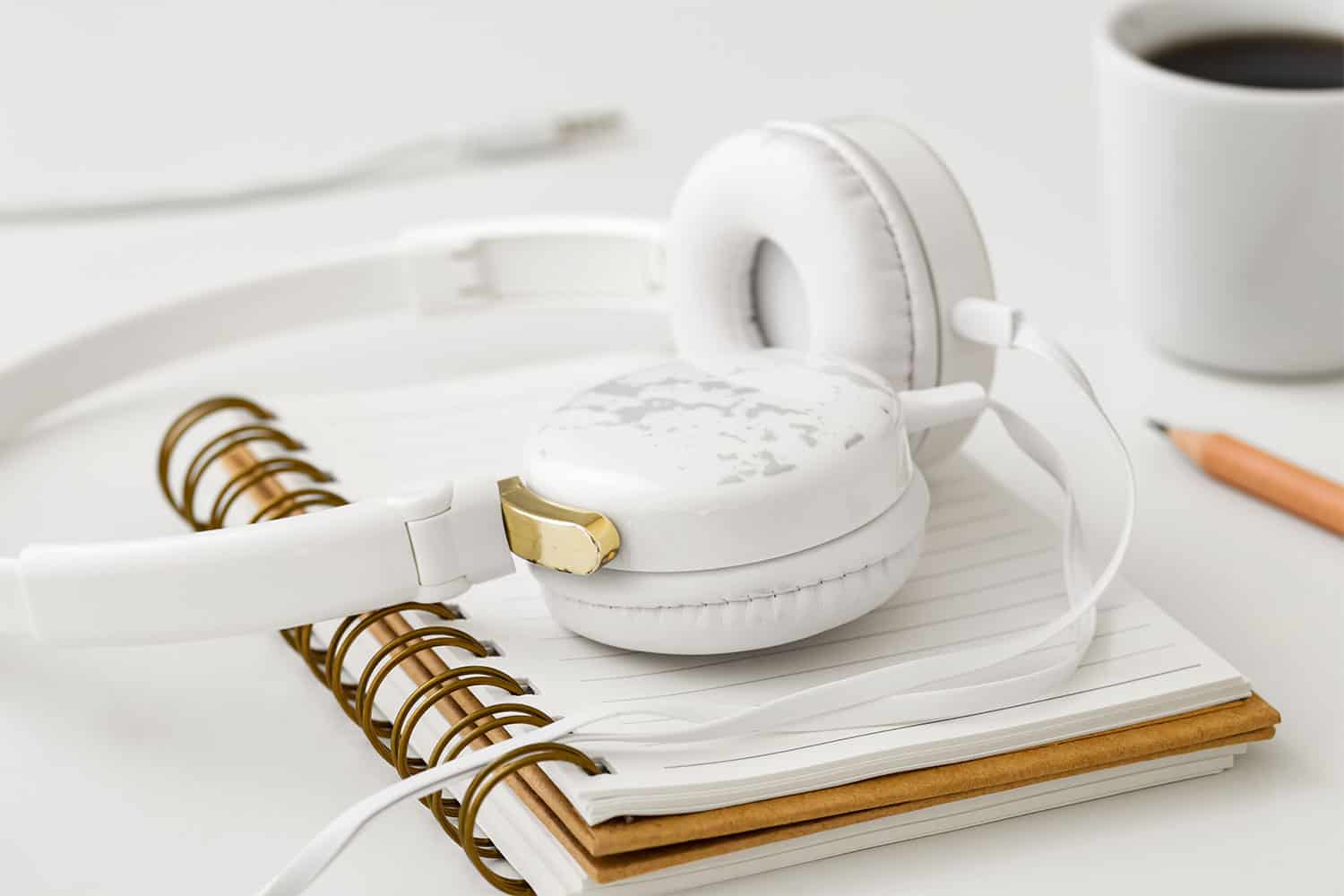 White over-ear headphones resting on a notebook with a gold paperclip, a cup of coffee in the background, and a pencil on a clean white desk, emphasizing music, technology, and workspace productivity.