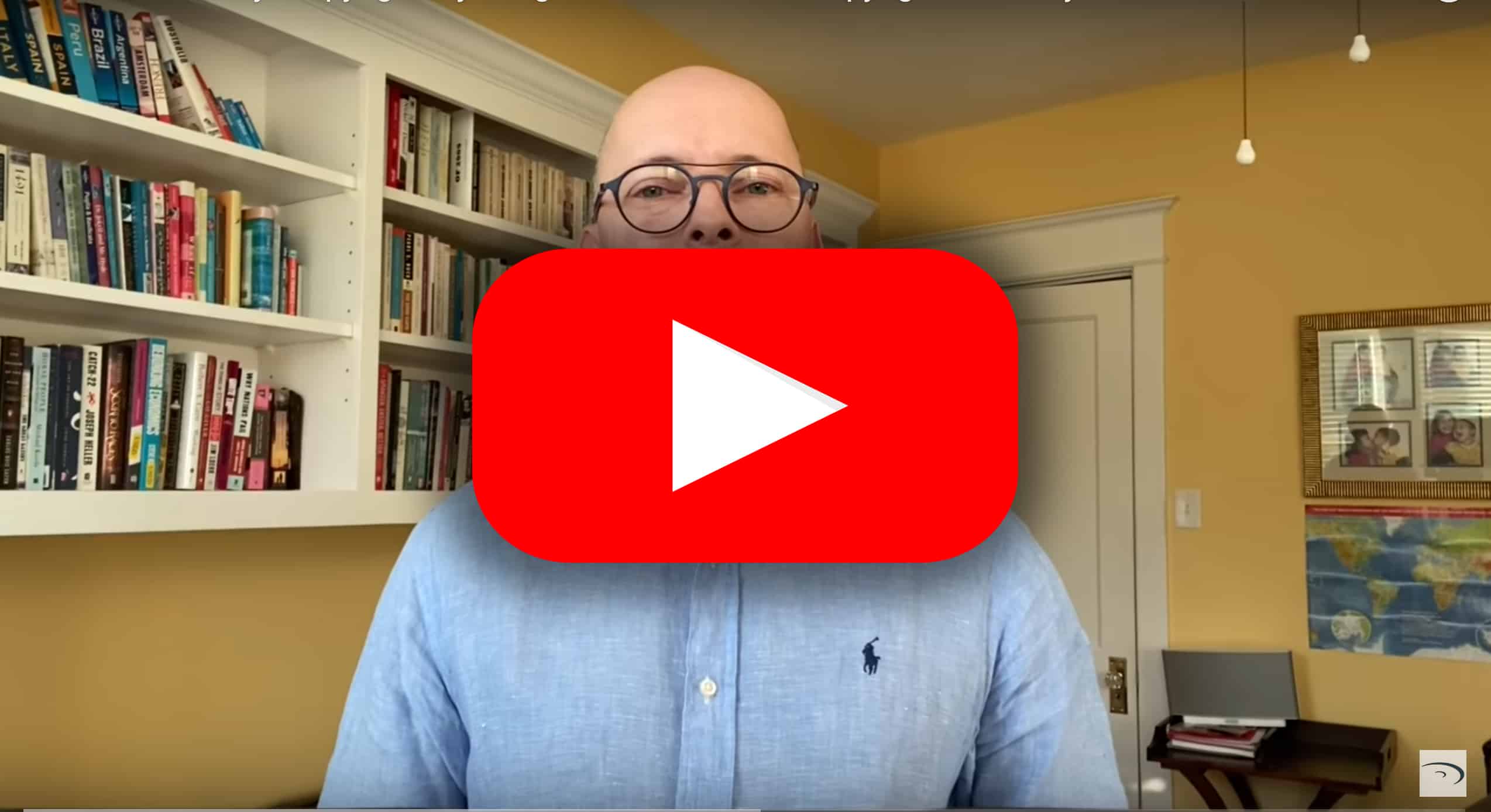 Professional man in blue shirt with glasses in home office or library setting, with bookshelves and framed photos in background.