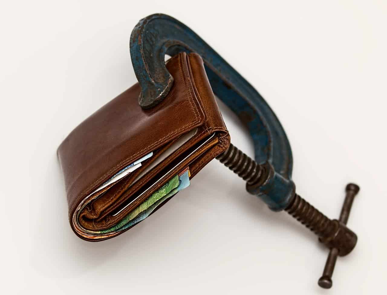 A vintage wallet clamped in a rusty C-clamp, with visible currency notes inside. The image symbolizes financial pressure, budget constraints, or the challenge of managing money.