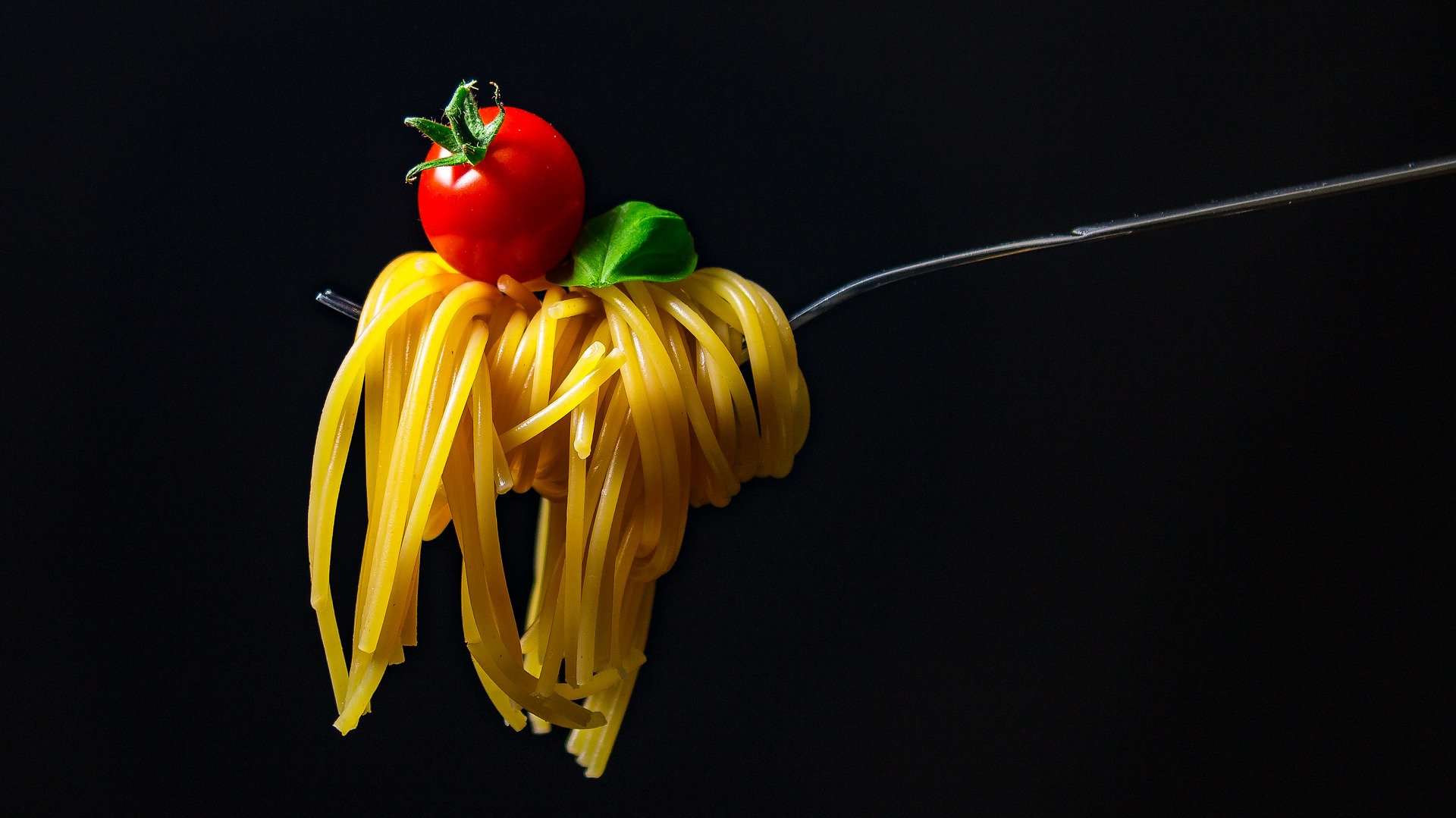 Fresh spaghetti noodles with a cherry tomato and basil leaf on a fork against a black background, emphasizing food presentation, Italian cuisine, and gourmet food photography.