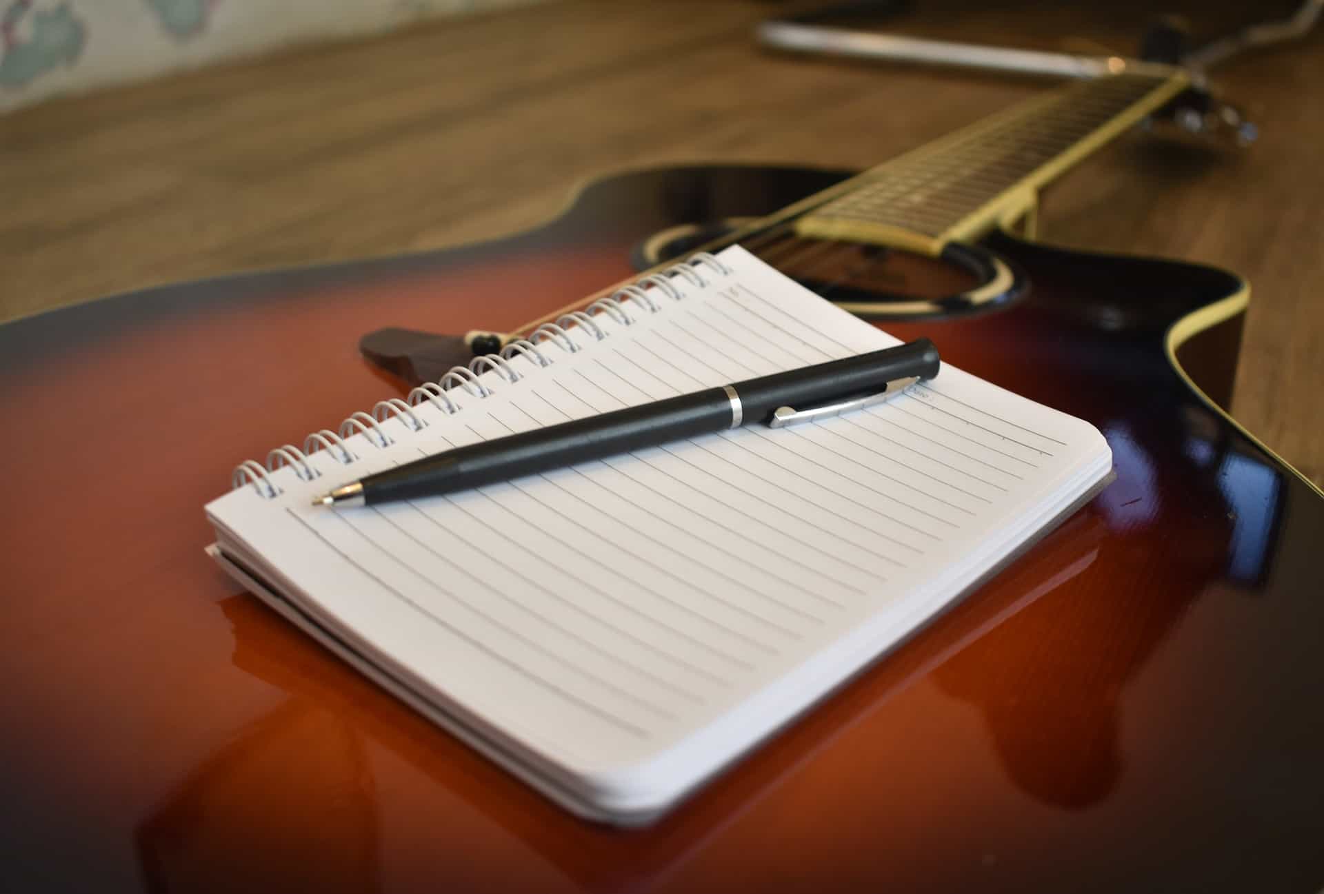 Aspirational image of a songwriting setup featuring an acoustic guitar, a spiral notebook for lyrics, and a black pen, perfect for musicians, songwriters, and music creators.