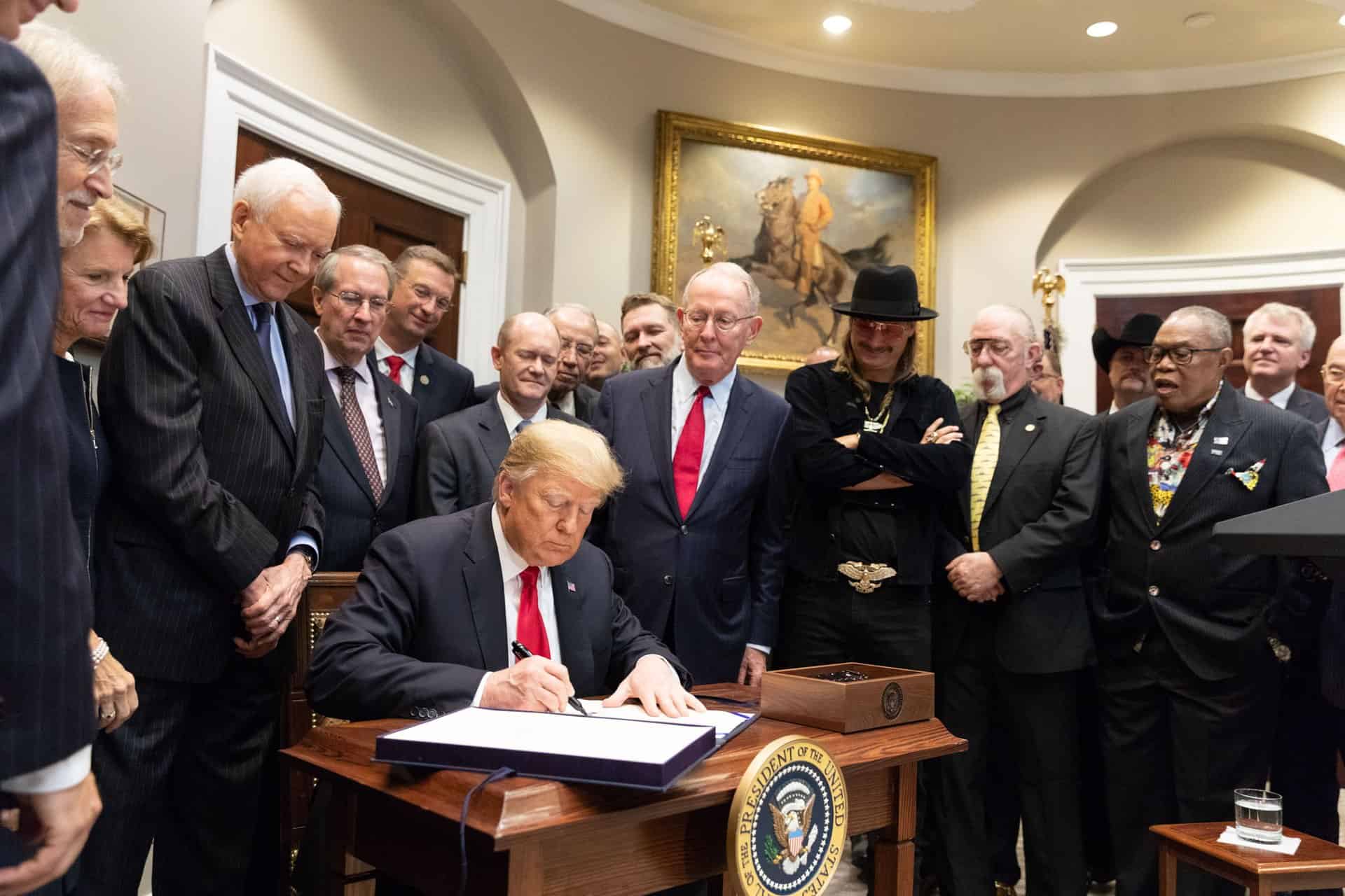 Donald Trump signing a document at a formal event with a group of men in suits and casual attire, some wearing accessories like hats and necklaces, in a well-decorated room with artwork and official decor.
