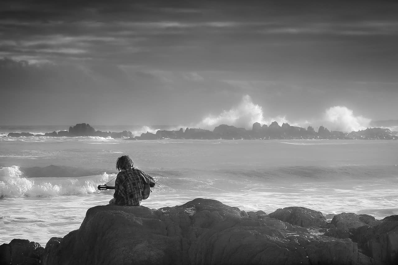 Boy playing guitar on rocks by the ocean at sunset, black and white photograph, coastal scenery, relaxing music, nature and music ambiance, serene seascape, artist on seaside, calming ocean waves, music and nature photography, Hypebot SEO focus relevance.