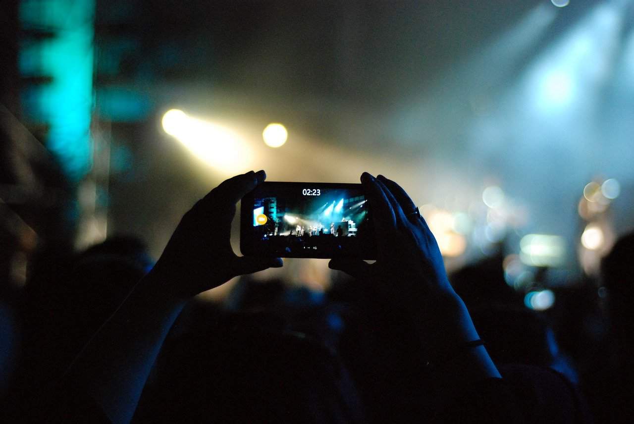Concert live music event captured with a smartphone in a dark setting with stage lighting and audience silhouette, vibrant blue and yellow hues, music performance photography, entertainment and concert photography, night music event photography.
