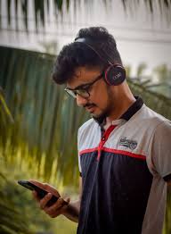 Headphone-wearing man using smartphone outdoors, listening to music or podcast in natural setting, vibrant greenery in background.