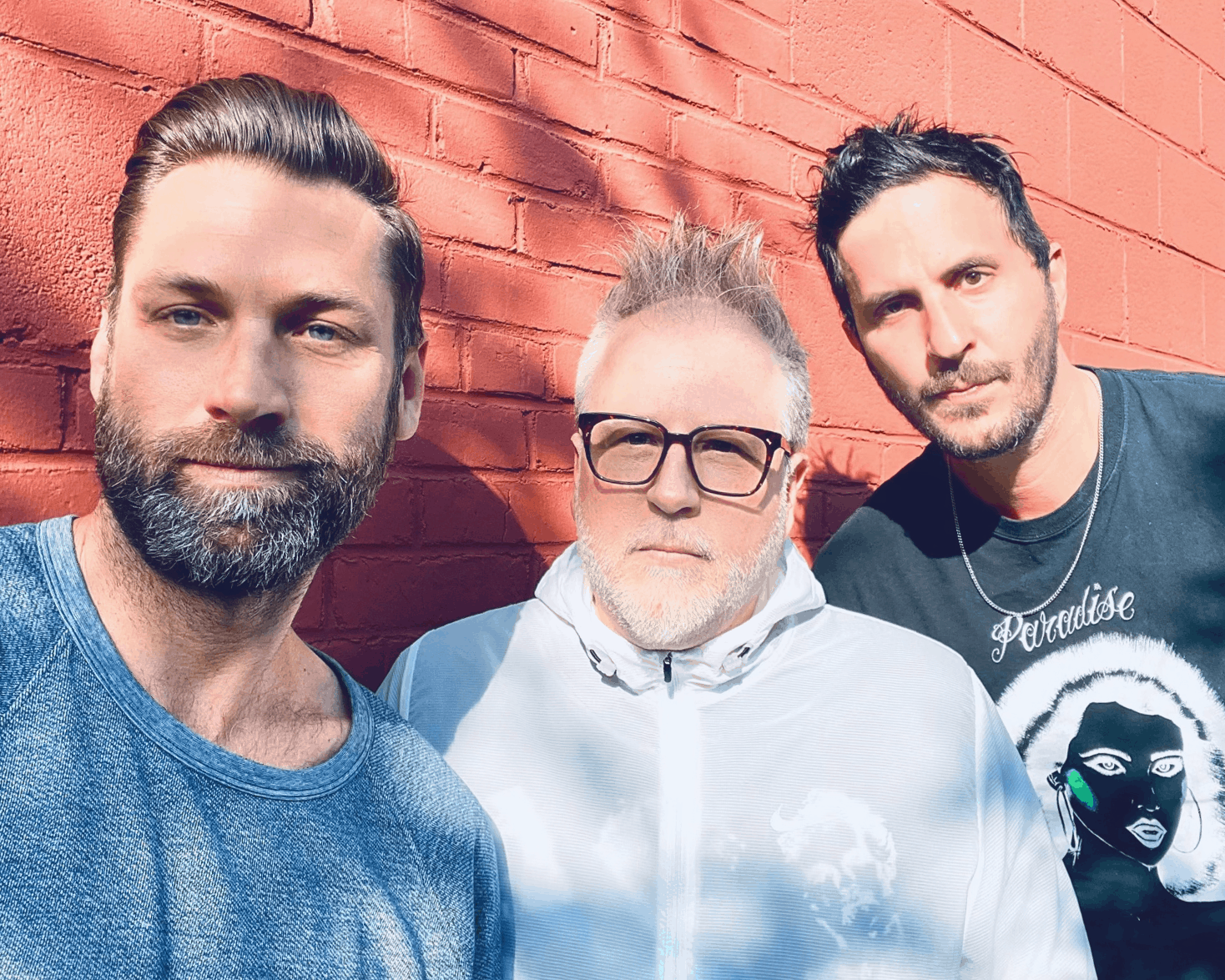 Bearded men standing against red brick wall, sunlight casting shadows, casual style, close-up, modern music influencers, urban background, community, artistic expression, Hypebot image.