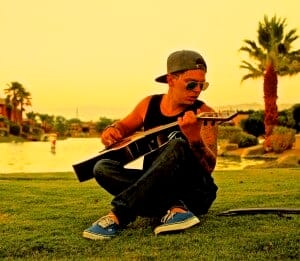 Guitar-playing young man sitting outdoors in a sunny park with palm trees, wearing sunglasses and a hat.