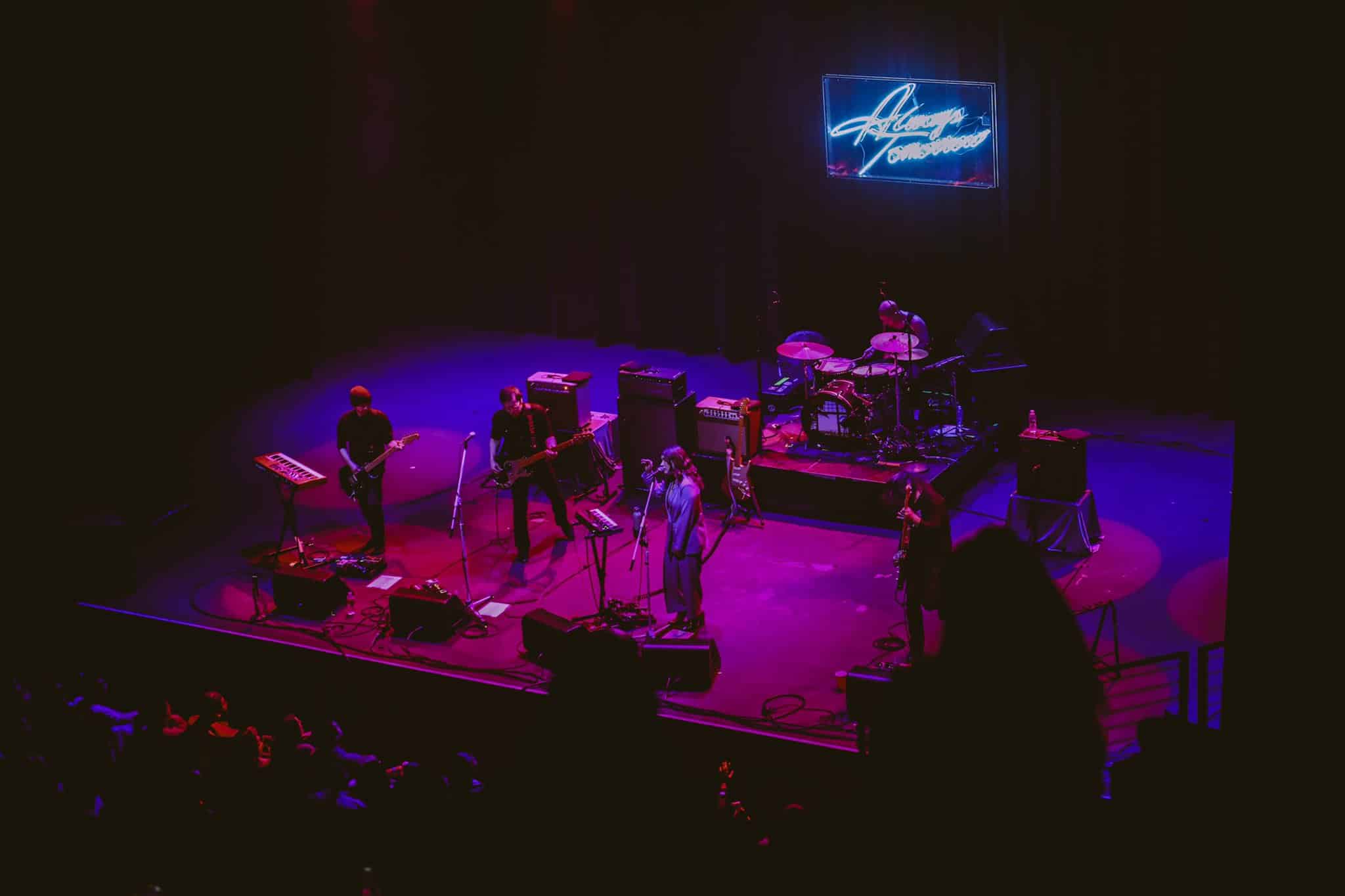 Live music performance on stage with band playing instruments under colorful lighting, neon sign in background, showcasing modern concert experience.