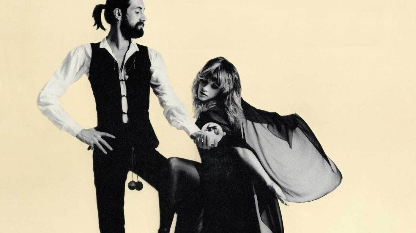 Bold black and white photograph of a stylish man with a ponytail and beard holding a woman's hand, both dressed in fashionable vintage attire, with a plain neutral background.