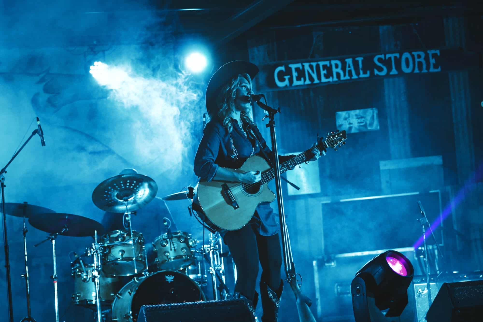 Guitarist performing live on stage at a music venue, blue lighting and atmospheric smoke, with a "General Store" sign in the background, showcasing a vibrant concert scene, music performance, live music, stage lighting.