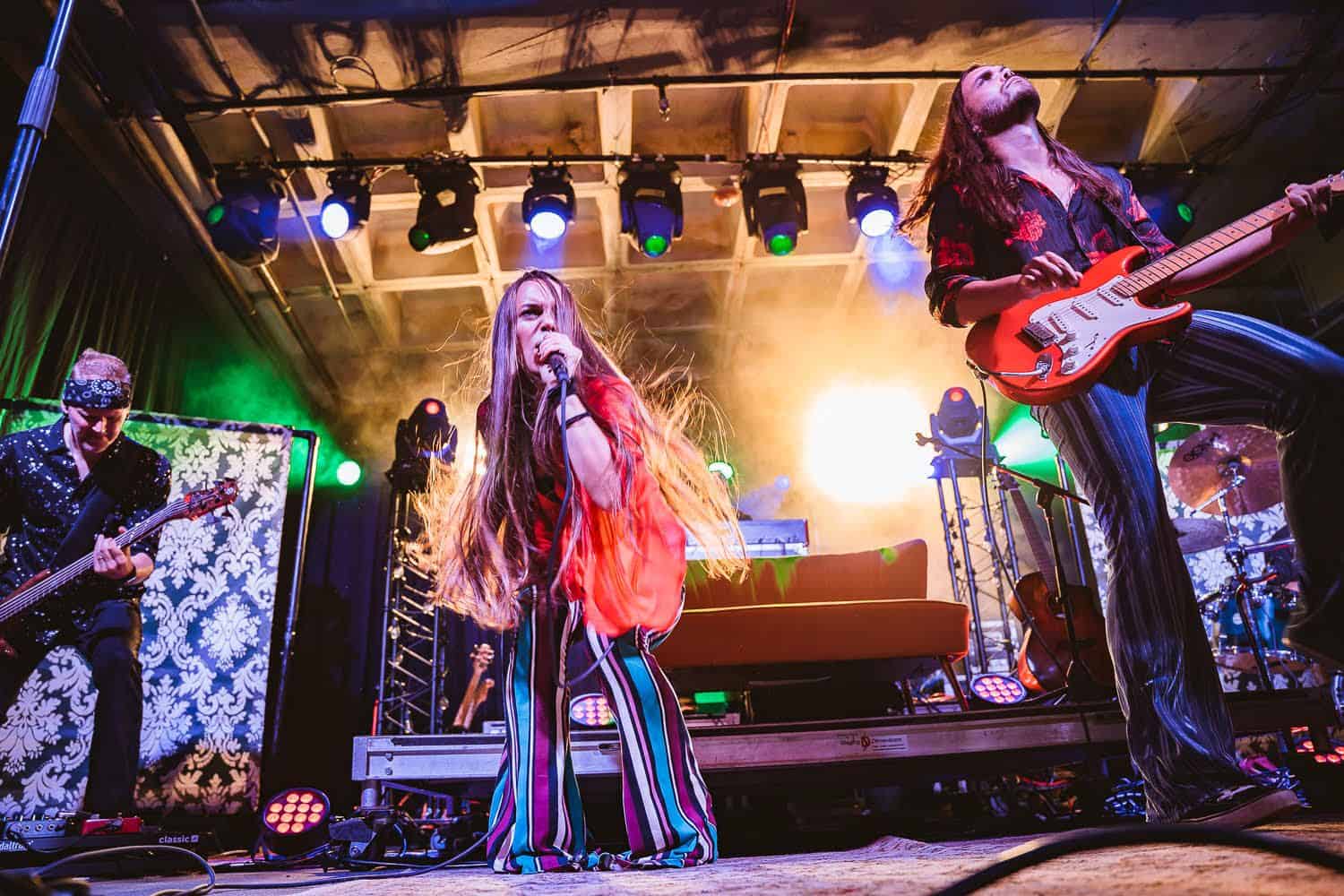 Vibrant live music performance featuring female lead singer and guitarists on stage with colorful lighting and dynamic stage setup, showcasing concert atmosphere and musical artistry.