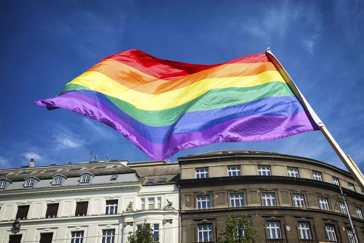 Vibrant rainbow pride flag flying high in front of historic city buildings against a clear blue sky, symbolizing LGBTQ+ pride, diversity, and inclusion.