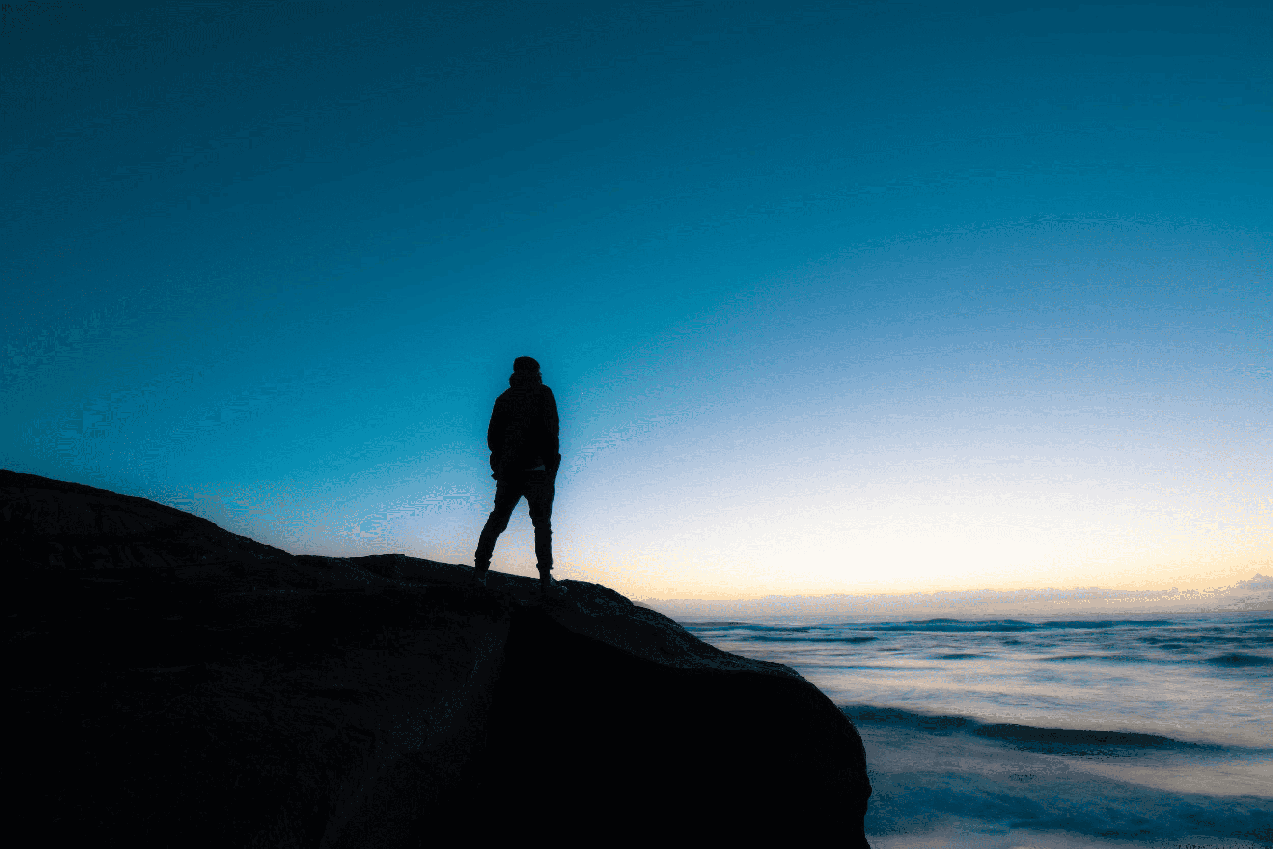 Silhouette of a person standing on a rocky cliff overlooking the ocean at sunset, creating a peaceful and reflective mood, ideal for nature and travel content.