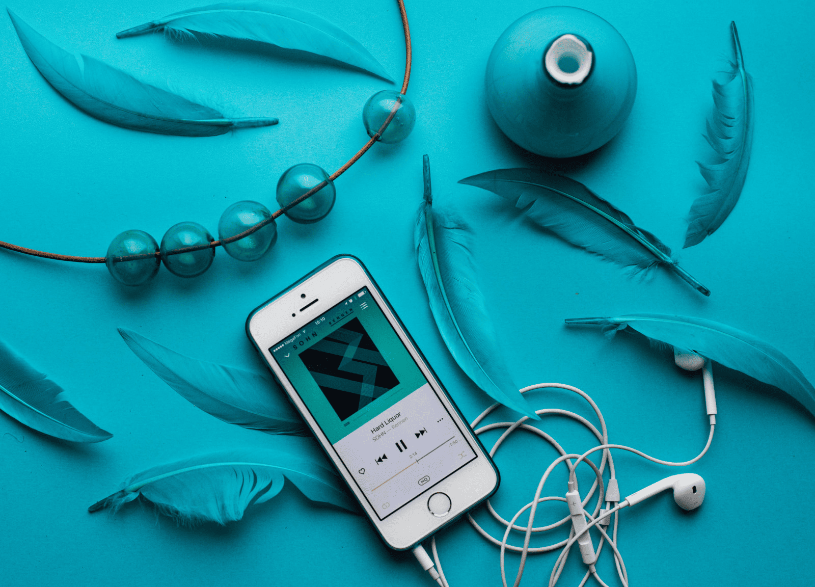 Futuristic music playlist on smartphone with white earphones, surrounded by blue feathers, glass beads, and a ceramic vase on vibrant turquoise background. Ideal for digital music trends and modern Spotify playlists.
