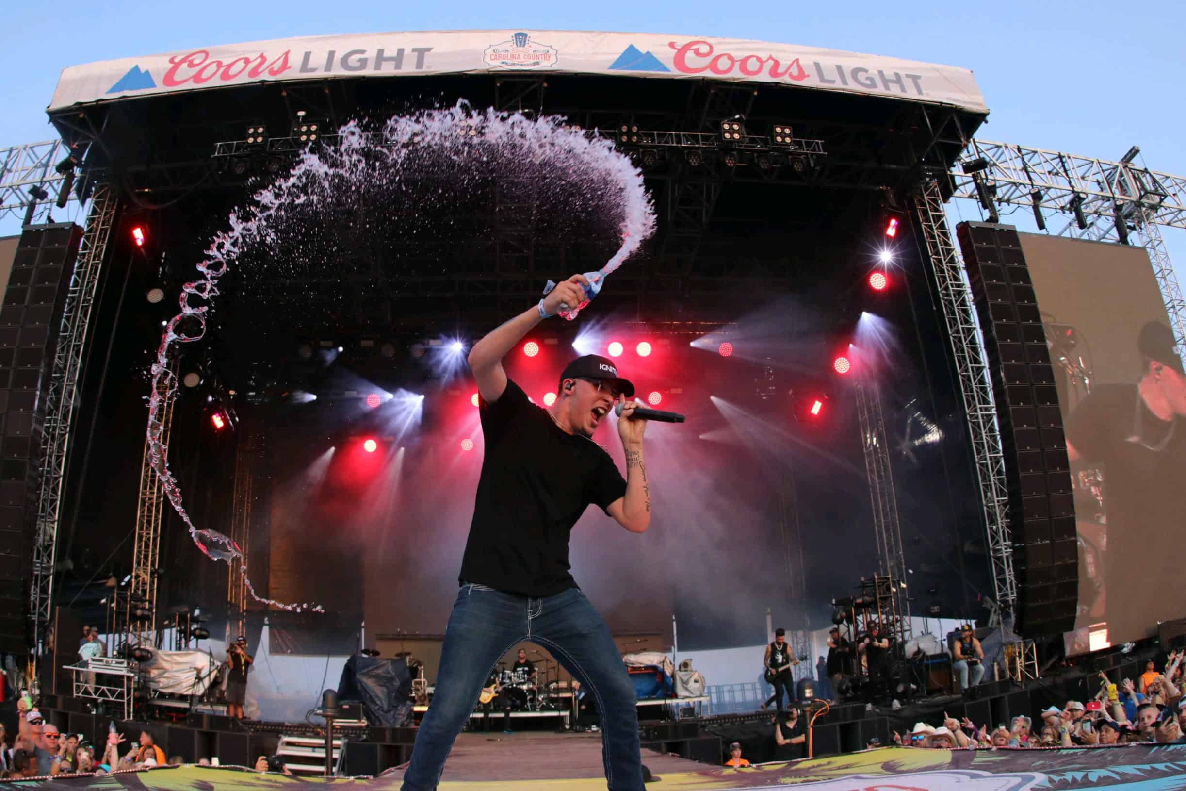 Energetic performer on stage at a music festival, holding a microphone and spraying water during a lively concert, with vibrant lighting and an engaged audience.