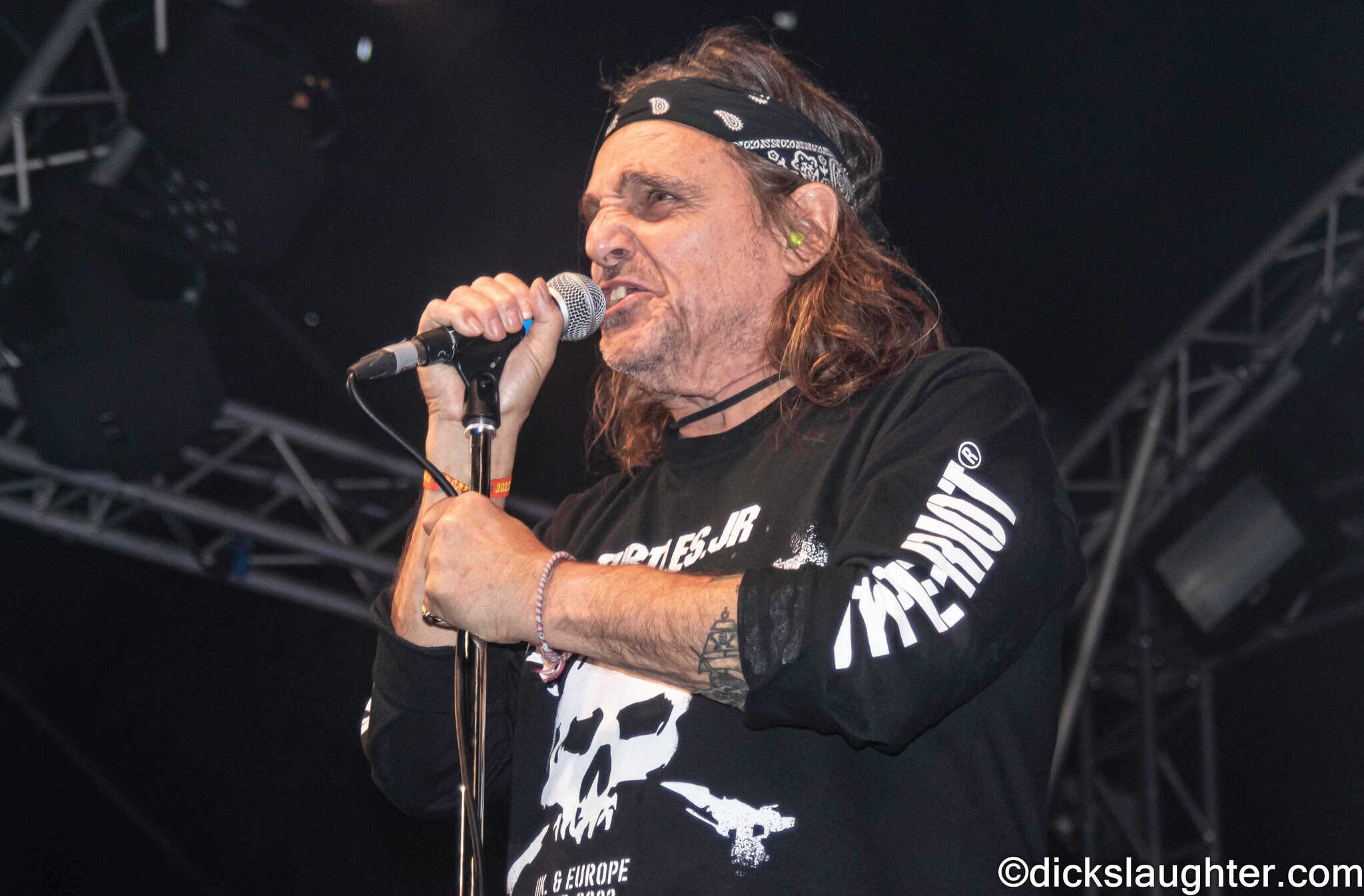 A rugged male singer performing live on stage, holding a microphone and wearing a bandana and black graphic sweatshirt, in an energetic music concert setting.