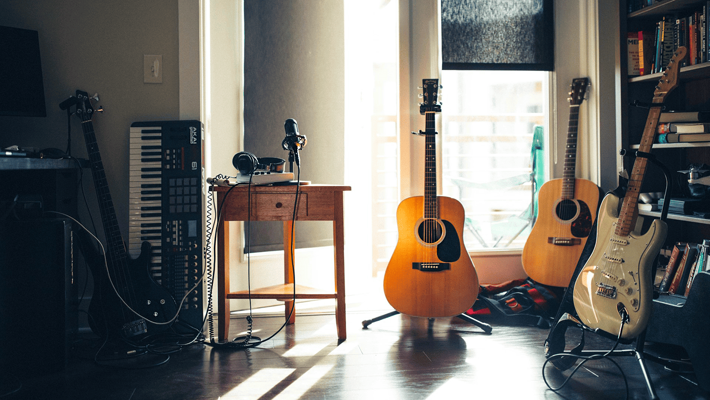 Guitar and music production setup in a home studio with sunlight streaming in, featuring acoustic and electric guitars, a MIDI keyboard, microphone, and other musical instruments.