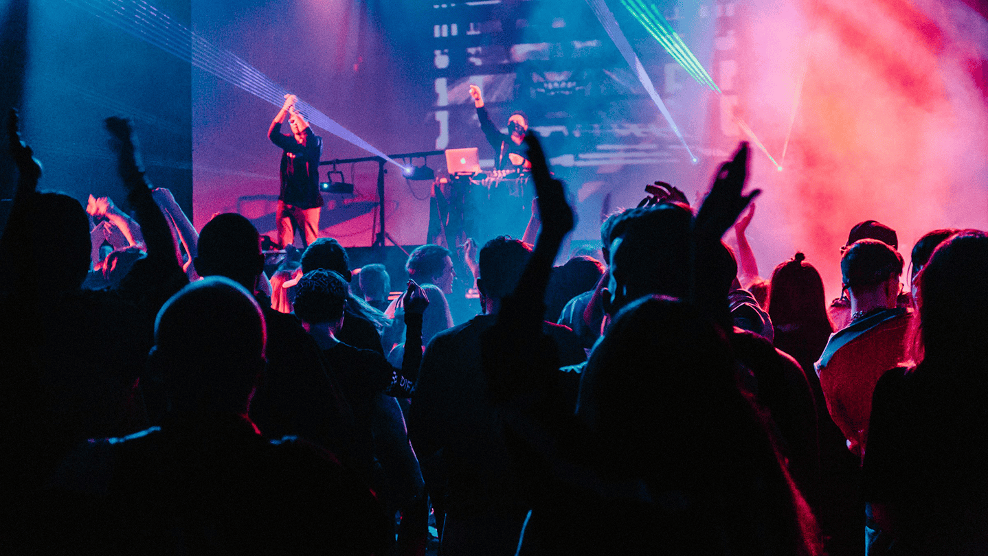 Electronic music concert with DJ performing on stage, colorful laser light effects, and an energetic crowd dancing and enjoying the live music experience.
