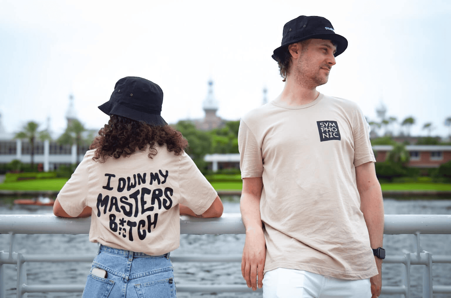 Down my Masters B*tch t-shirt, young woman and man wearing hats by waterfront with city skyline and park in background, casual streetwear style, trendy fashion, youth culture, relaxed vibe, outdoor recreational scene, urban lifestyle.