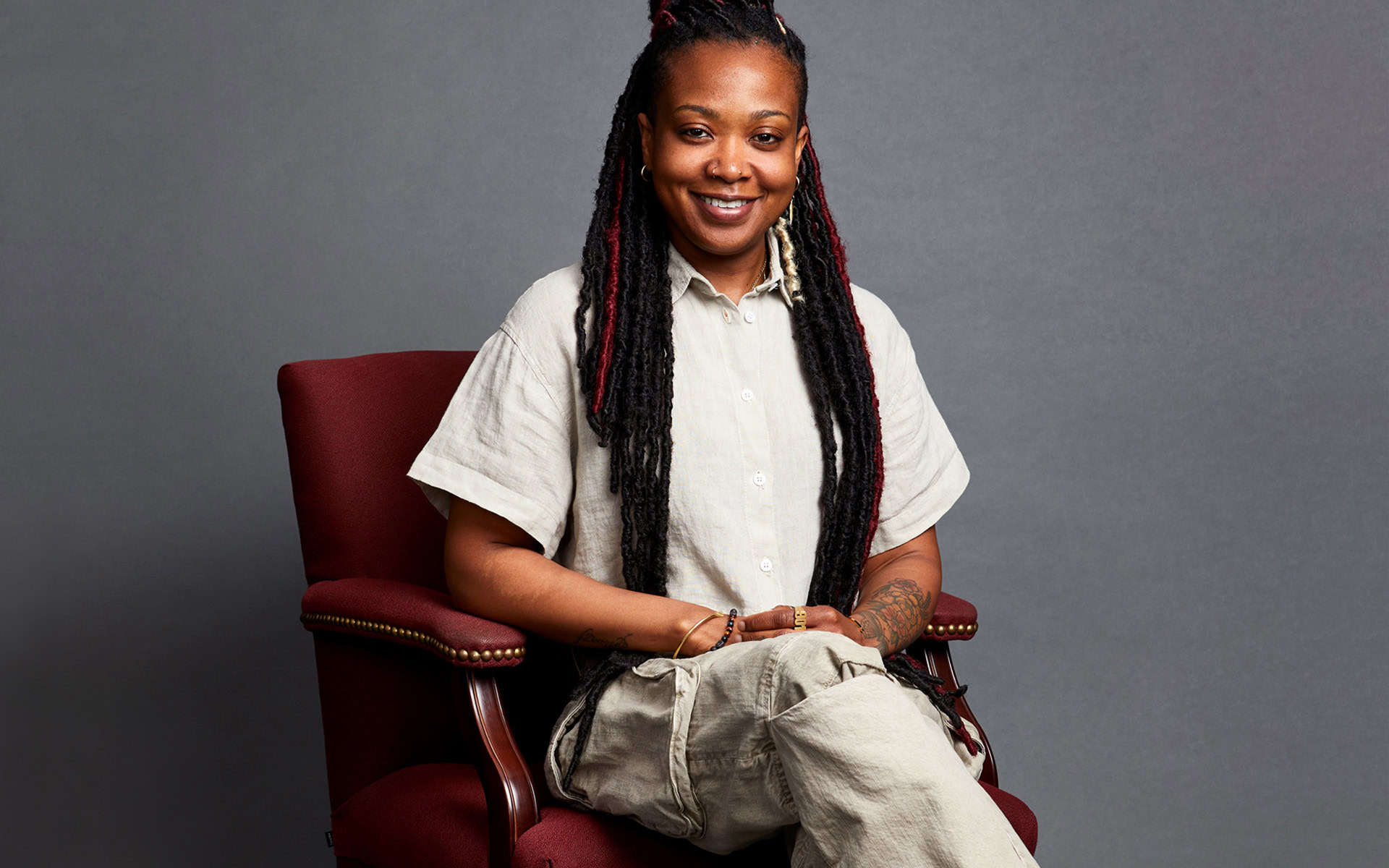 Vibrant Black woman with dreadlocks, smiling confidently, seated on a stylish red armchair against a neutral gray background, embodying creativity and empowerment.