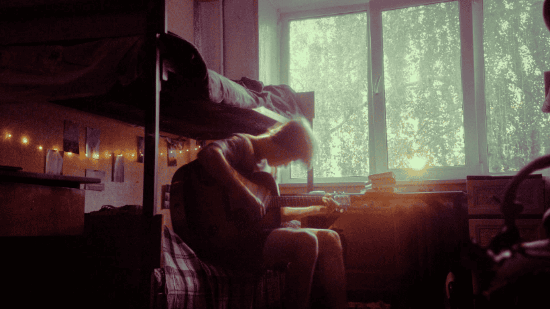 Guitarist playing acoustic guitar in a cozy, sunlit dorm room decorated with fairy lights and pictures, creating an inspiring environment for musicians and music lovers.