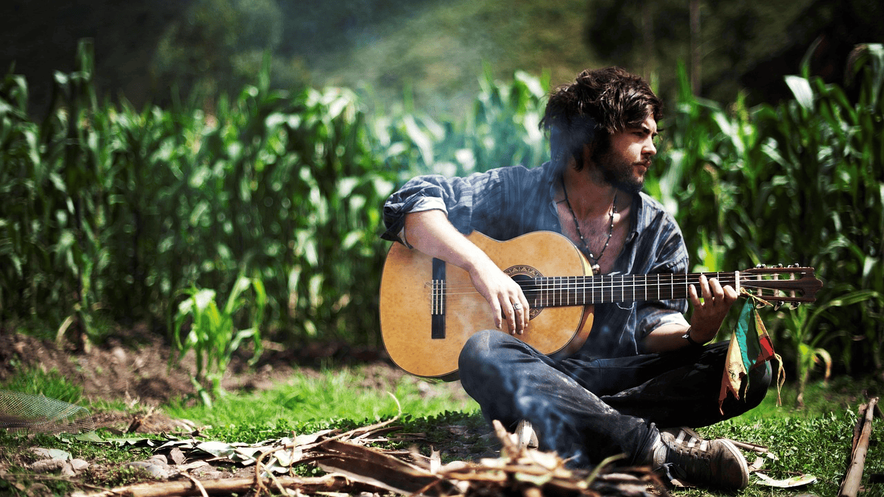 A man playing acoustic guitar outdoors in a lush green field, enjoying nature, with sunlight filtering through the trees.
