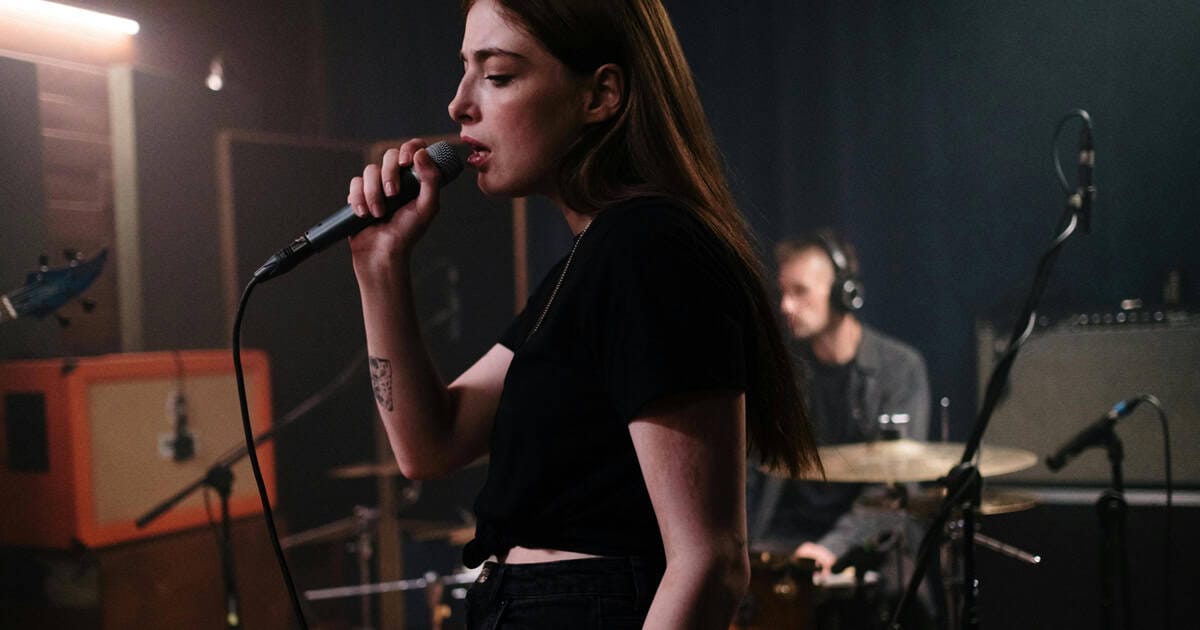 Vocalist singing into microphone in recording studio with band members and musical equipment, capturing a live music performance or rehearsal.