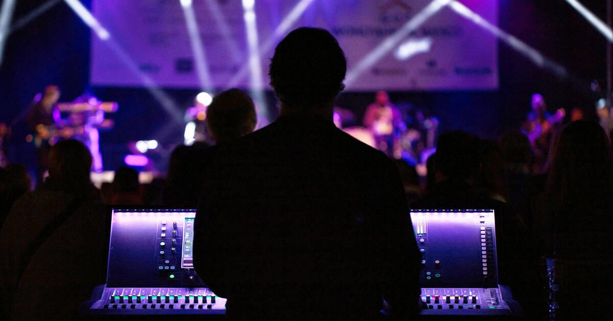 Live concert sound engineer working with digital audio mixing console at music event, vibrant stage lighting, concert audience, music performance, event production, live sound, professional audio setup, event management.